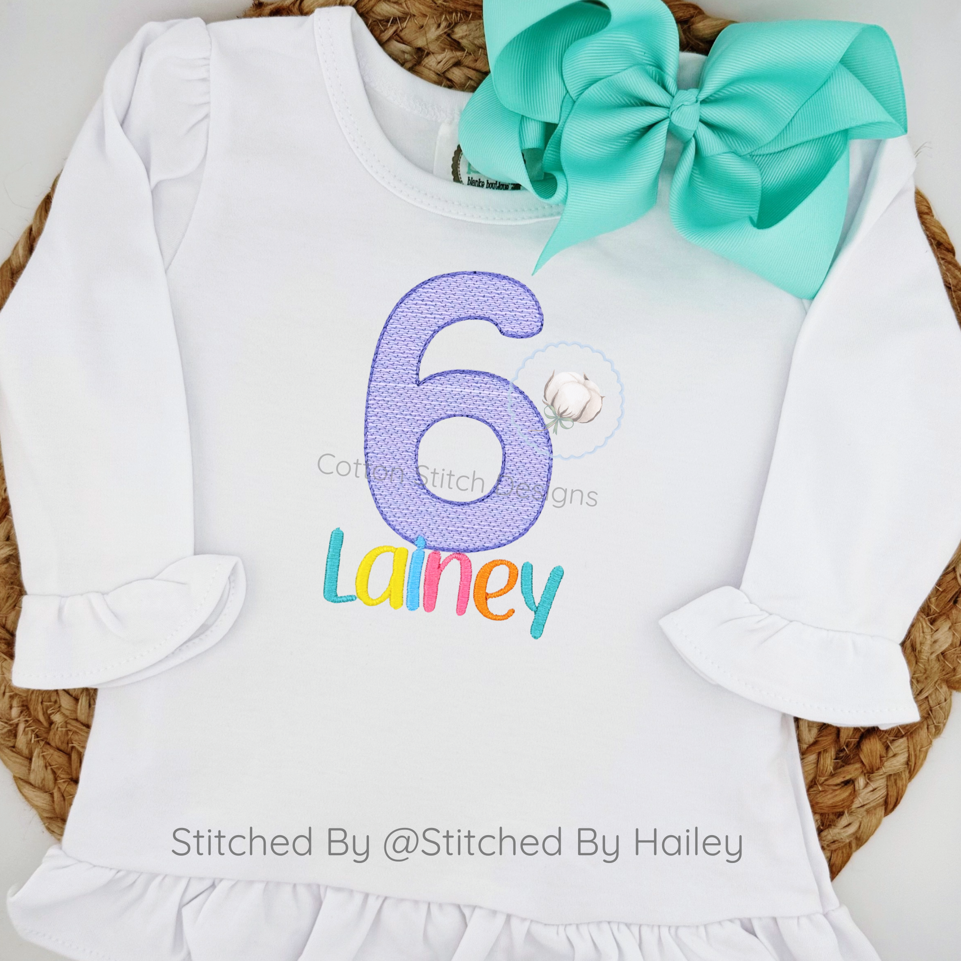 Machine Embroidery design Cotton Stitch Designs White dress with number 6 and name 'Lainey', paired with a large teal bow, on a woven surface.
